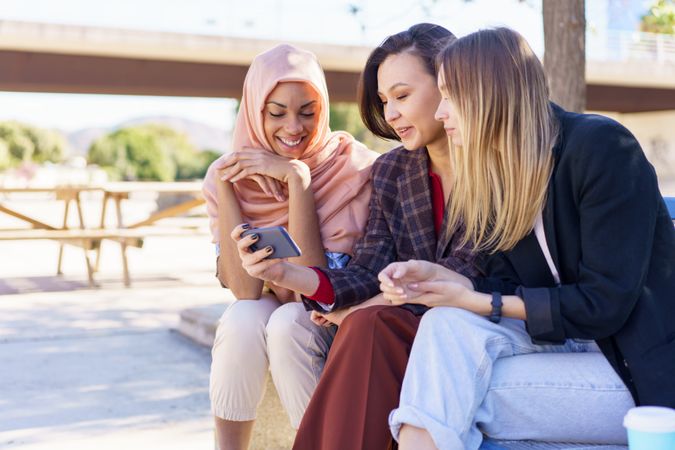 Three women sitting on outdoor park bench watching smartphone