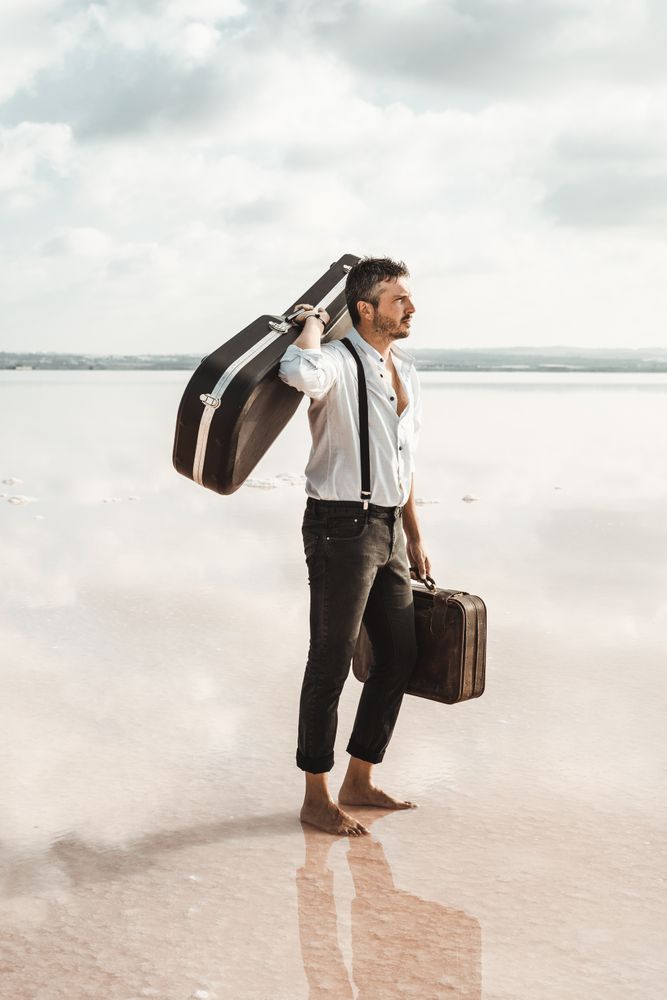 Focused stylish man with suitcase and guitar gig bag on seaside
