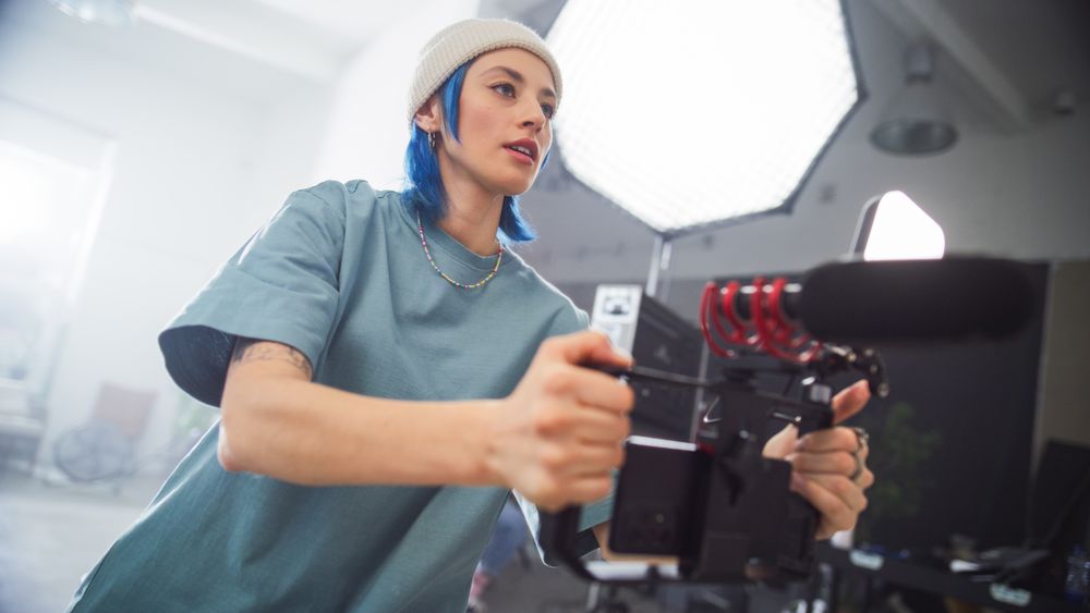 Focused Young Female Videographer With Blue Hair Adjusts Her Camera During An Indoor Film Shoot, Capturing The Essence Of Modern Filmmaking And Creative Production In A Professional Setting.