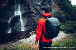 Back view of a man in red jacket with backpack standing facing waterfall during daytime 0LJwX4