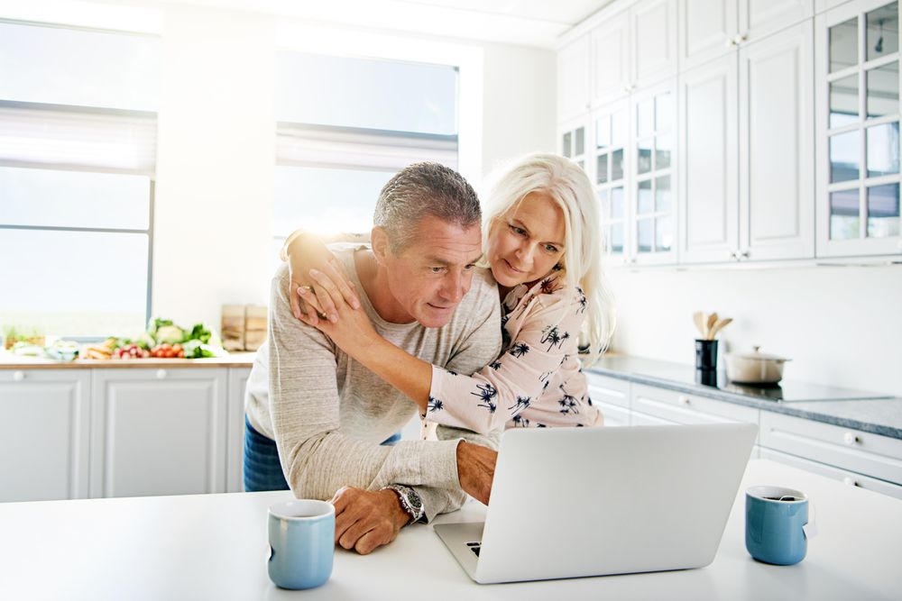 Grey haired couple embracing in the the kitchen with a laptop and cups of tea