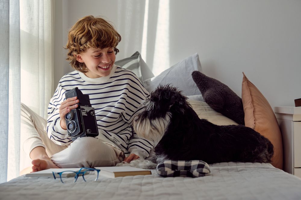 Glad schoolboy with retro photo camera and dog on bed