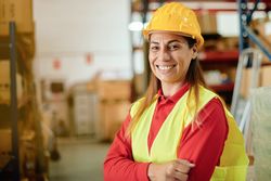 Portrait of a happy adult working woman looking at the camera inside a warehouse wearing a hard hat and safety clothing - Focus on the Face