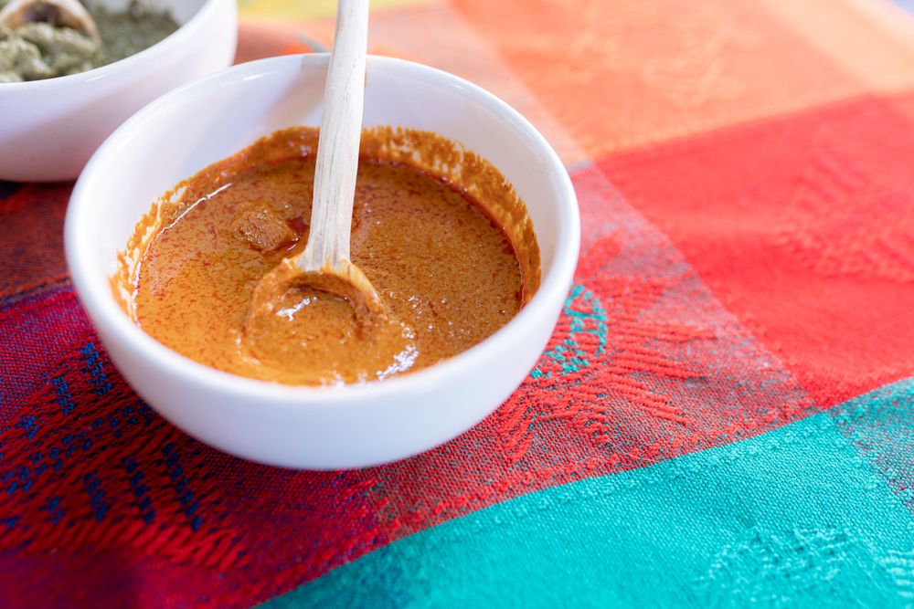 A bowl of yellow mole sauce with a wooden spoon on top of a colorful traditional tablecloth