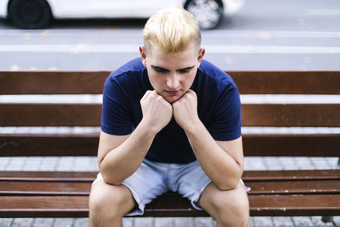 Young sad man sitting on a bench in the street with his hands on his worried face