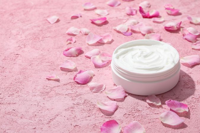 Jar with cream and rose petals on pink background, space for text