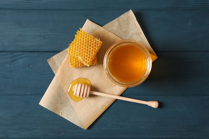 Glass jar with honey, dipper, honeycombs and parchment on wooden background, copy space