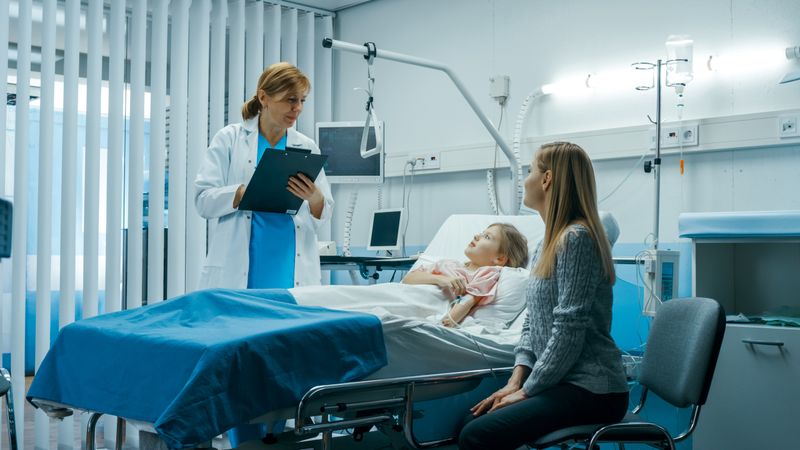 Sick Little Girl Lies on a Bed In the Hospital, Friendly Doctor With Clipboard Asks Where it Hurts, Mother Sits Beside Bed