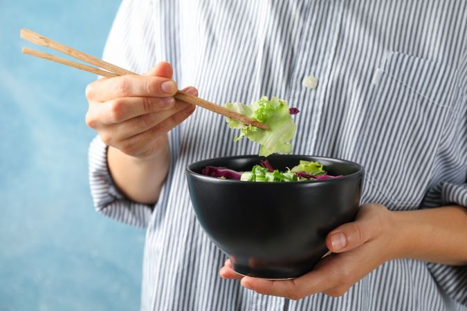Young woman eat salad with chopsticks on blue background, space for text