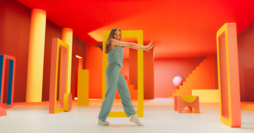 Portrait of White Stylish Female Dancer Performing in a Funky Colorful Decorated Studio While Looking at Camera. Woman Dancing in Abstract Environment, Jumping Through Geometric Shapes