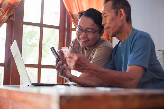Happy adult son and older father pay online using laptop and credit card