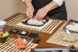 Chef in apron filling Japanese sushi rolls with rice in a nori sheet on a traditional mat 43XOOb