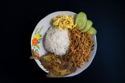 Flat lay of a plate with rice, fried fish, fried noodles, sliced omelette, and cucumber on a dark background
