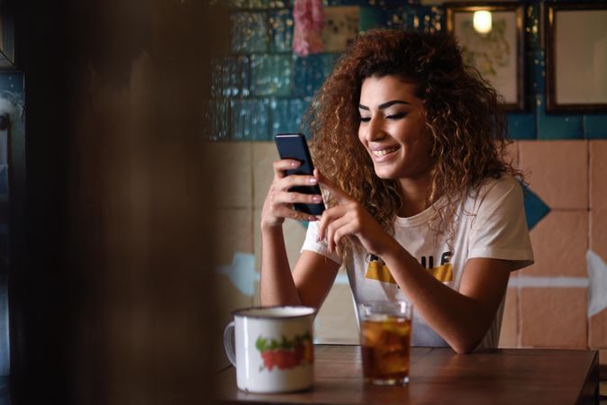 Arab woman in casual clothes smiling at her phone at restaurant table