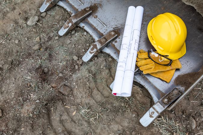 Technical Blueprints, Hardhat, Gloves and Protective Glasses Resting on Bulldozer Bucket Abstract