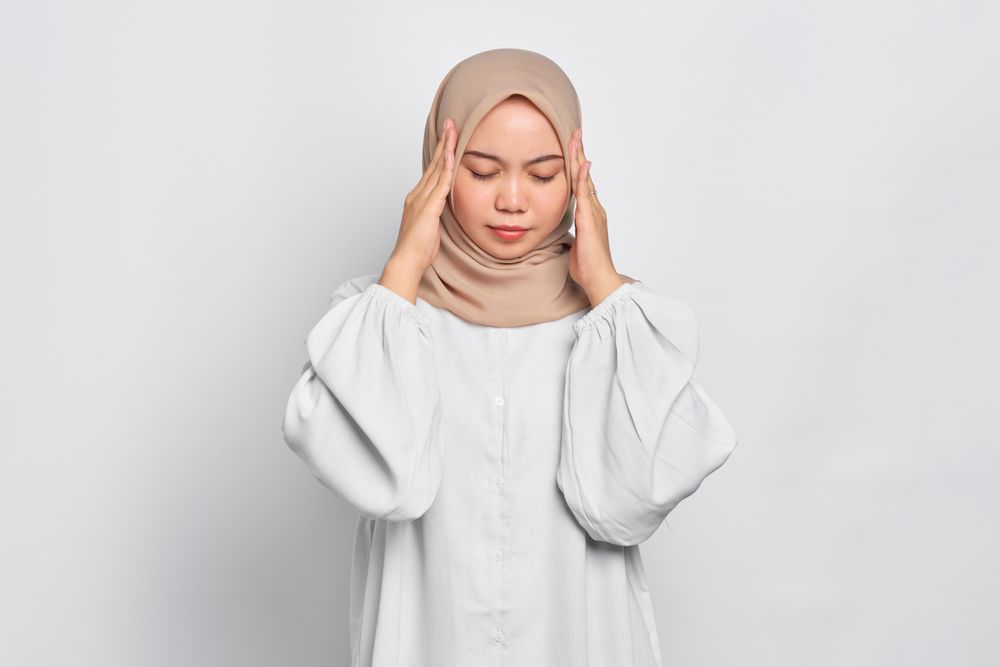 Stressed Asian female in headscarf with hands soothing her headache