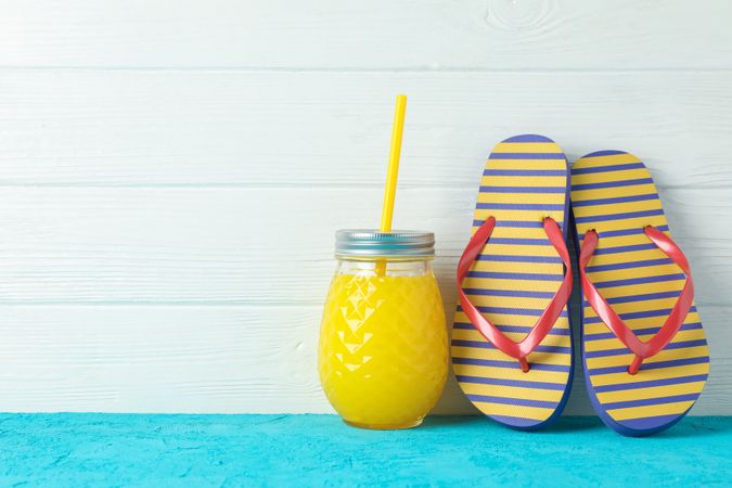 Flip flops and glass jar with fresh juice on color table against wooden background, space for text. Summer vacation backdrop