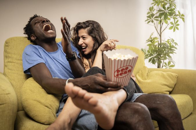 Exuberant diverse couple laughing out loud while watching a comedy movie at home on the sofa.