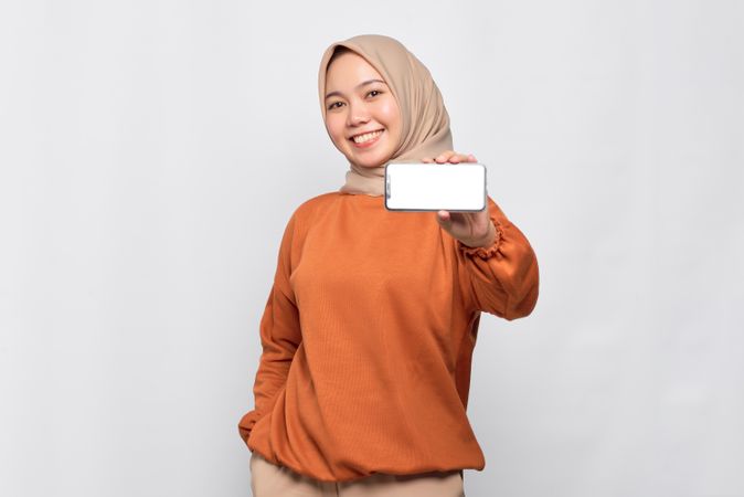 Confident Muslim woman smiling while holding up smart phone with mock up screen