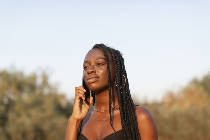 A young, Black woman with braids in her hair, happily talks on her smartphone during a sunset stroll