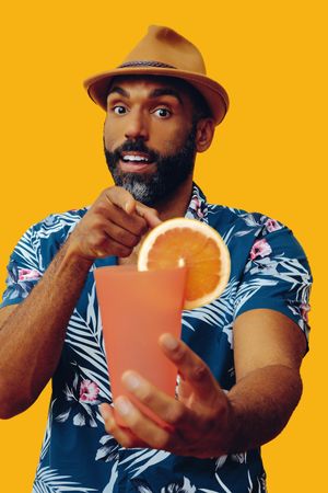 Male passing a cocktail and pointing at camera while wearing colorful shirt with yellow background