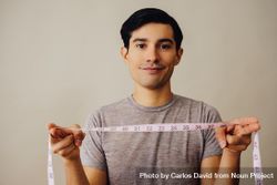 Hispanic male holding measuring tape between hands in beige studio shoot and looking at camera 5rjKP5