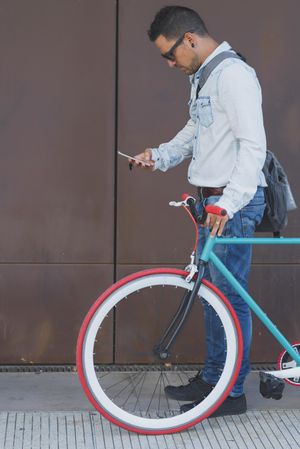 Male in sunglasses standing with red and green bicycle looking down at cell phone, vertical
