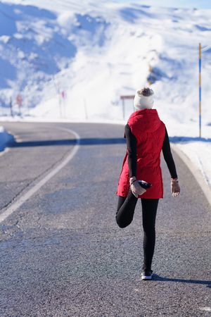 Healthy woman in wintry gear stretching quadriceps in the mountains on cold day