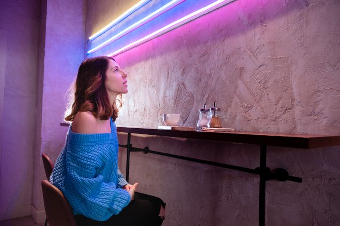 Positive woman with cup at table near wall with neon lamps