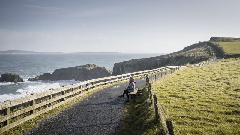 Woman sitting on bench on country road between sea and field