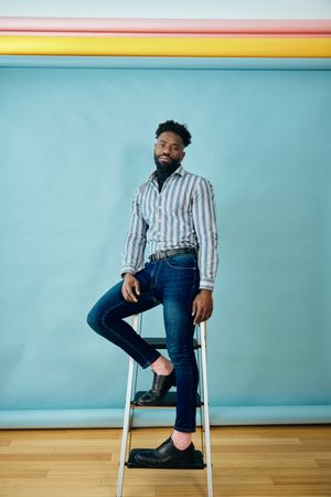 Black male model posing in blue studio in jeans and striped shirt on step ladder