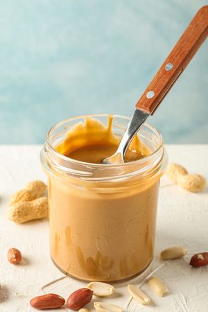 Glass jar with creamy peanut butter and spoon, and peanut on table against light background, space for text