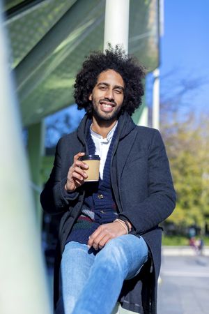 Front view of smiling Black man in jacket with warm beverage balancing between poles outside