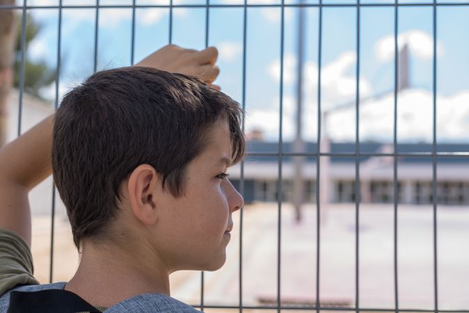 Boy looking to side while holding gate looking into school yard
