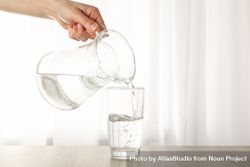 Hand pouring water in glass from pitcher in bright room with curtain bxkEZ4