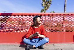 Happy woman in red sitting cross legged using a mobile phone outdoors on a bright day
