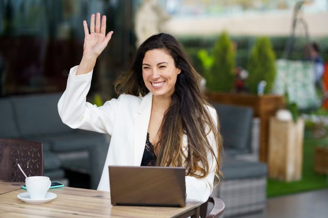 A Woman Enthusiastically Waving While Engaging in Remote Work at a Contemporary Outdoor Cafe