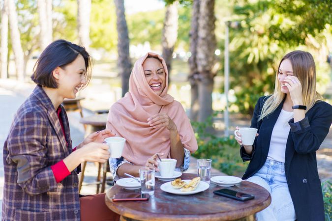 Three women chatting and laughing over coffee and baked goods on sunny outside table