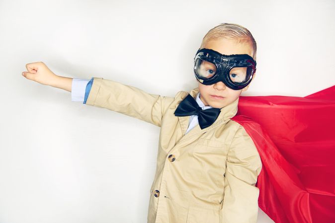 Serious blond boy wearing airplane goggles and cape with arm out