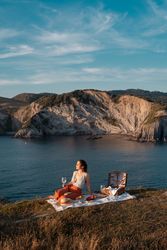Woman with glass of drink on picnic mat looking at sea and mountains