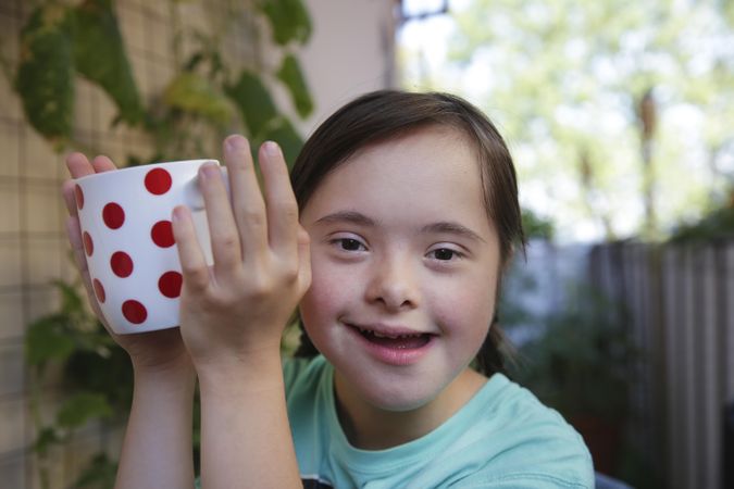 Little girl holding a white cup with red polka dots smiling at camera in backyard