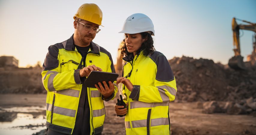 White Male Civil Engineer Talking To Hispanic Female Inspector And Using Tablet Computer On Construction Site of a New Building. Real Estate Developers Discussing Business, Excavators Working.