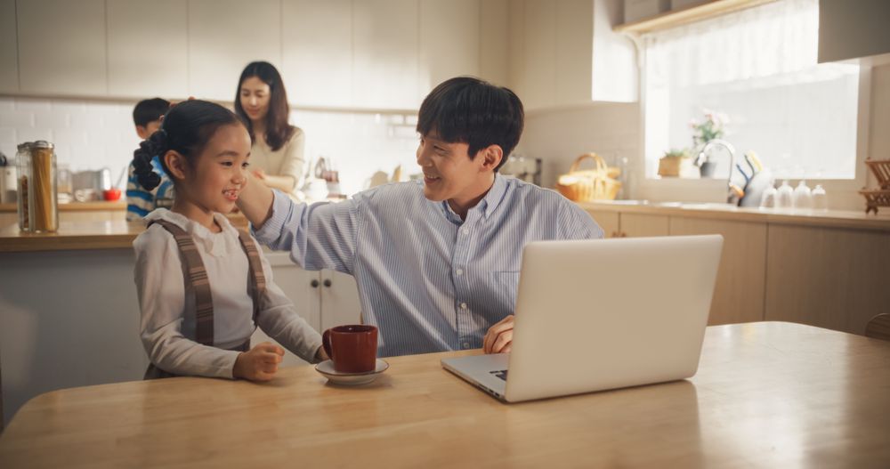 Portrait of Young Korean Father Using Laptop To Work from Home While his Wife and Kids are Working Together to Prepare Lunch.