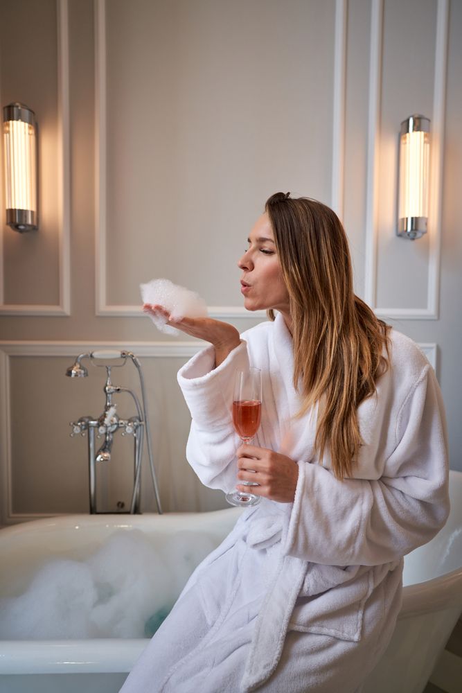 Woman in bathrobe with champagne glass sitting on bathtub edge