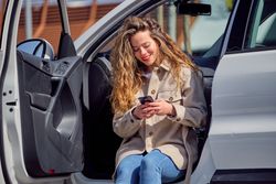 Smiling woman in coat browsing smartphone and sitting in car