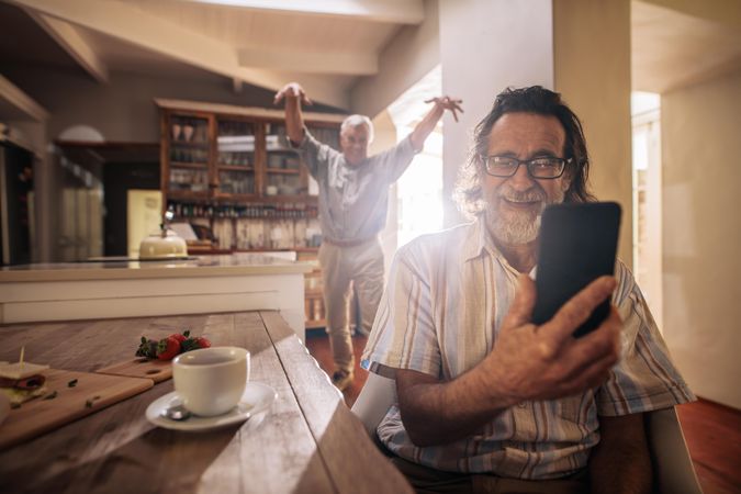 Happy older man taking selfie with friend standing at back making funny gestures