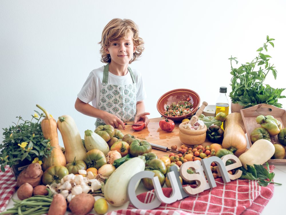 Adorable boy cutting tomato on chopping board near assorted veggies