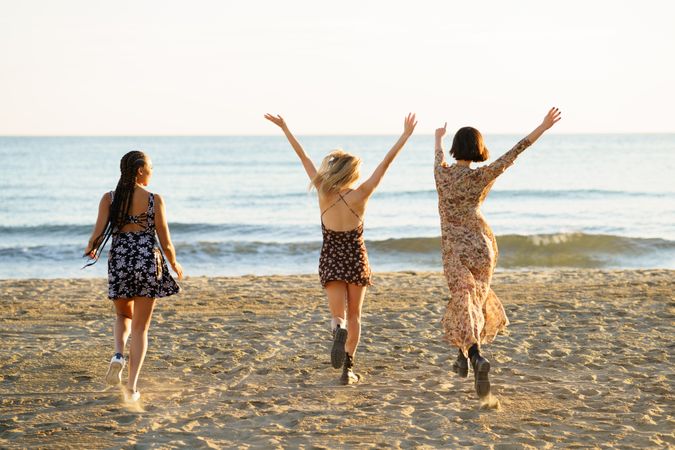 Backs of three happy women skipping towards the coast on beach with arms up