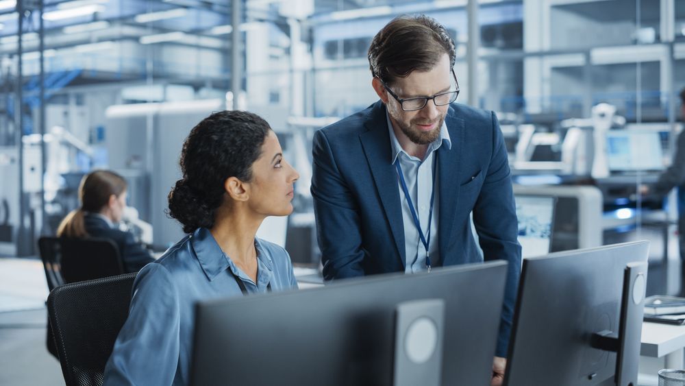 Portrait of a Young Male Team Leader and Hispanic Female Engineer Collaborating on a Project, Using Computer, Talking in a Factory with Robotic Arms Producing Devices for Tech Industries.