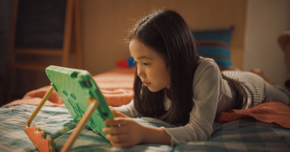 Smart South Korean Girl Playing Learning Puzzle Video Game on a Tablet Computer at Home in Her Children's Bedroom.